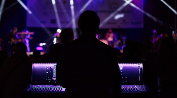 A man stands behind a digital audio console at a concert with purple lighting in the background